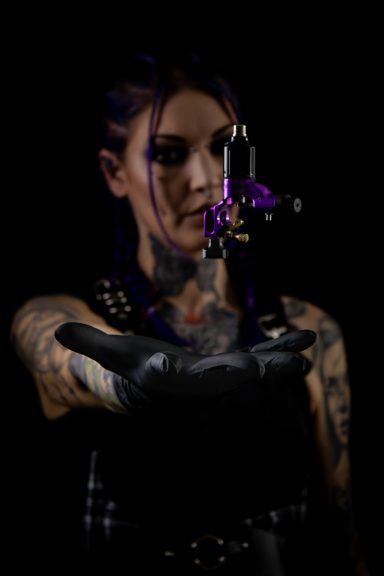 Femme tatoueuse portant des gants, tenant une machine à tatouer avec des lumières violettes.