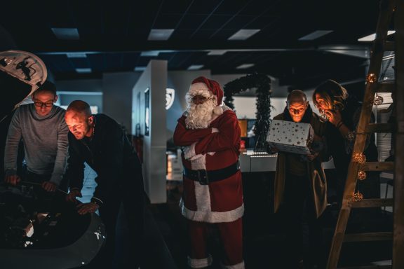 Un Père Noël observe un groupe de personnes près d'une voiture ouverte dans un atelier.