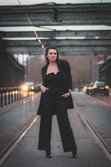 Femme en tenue noire posant sur une voie ferrée, avec un ciel nuageux en arrière-plan.