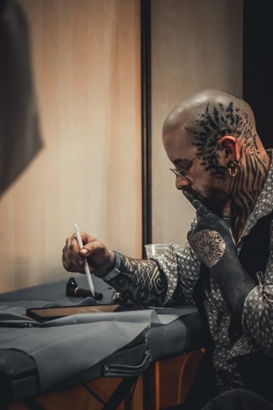 Homme tatoué assis à une table, observant attentivement un stylo et un carnet.