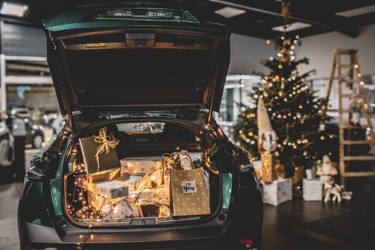 Coffre de voiture rempli de cadeaux et décoré de lumières, sapin de Noël en arrière-plan.